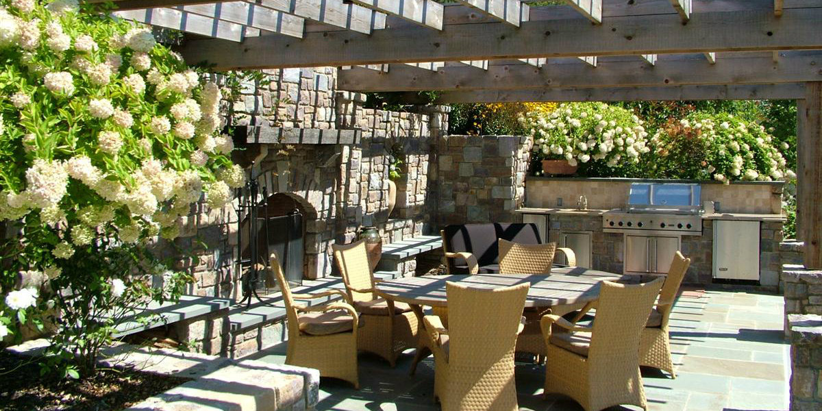 An outdoor patio with a pergola for shade has ample space for an outdoor seating arrangement and a custom outdoor kitchen.